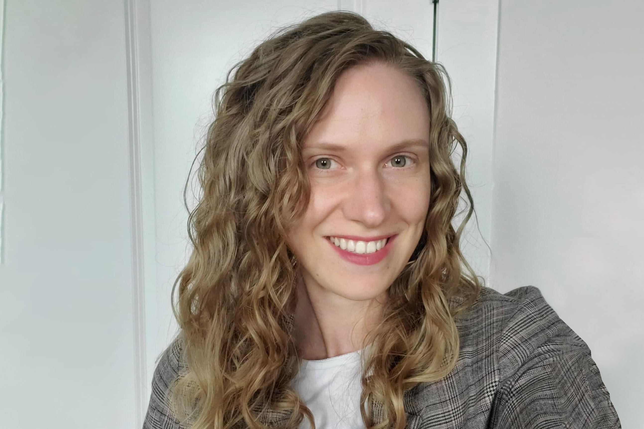 headshot of curly haired blonde woman in gray herringbone suit and white tee against a white background